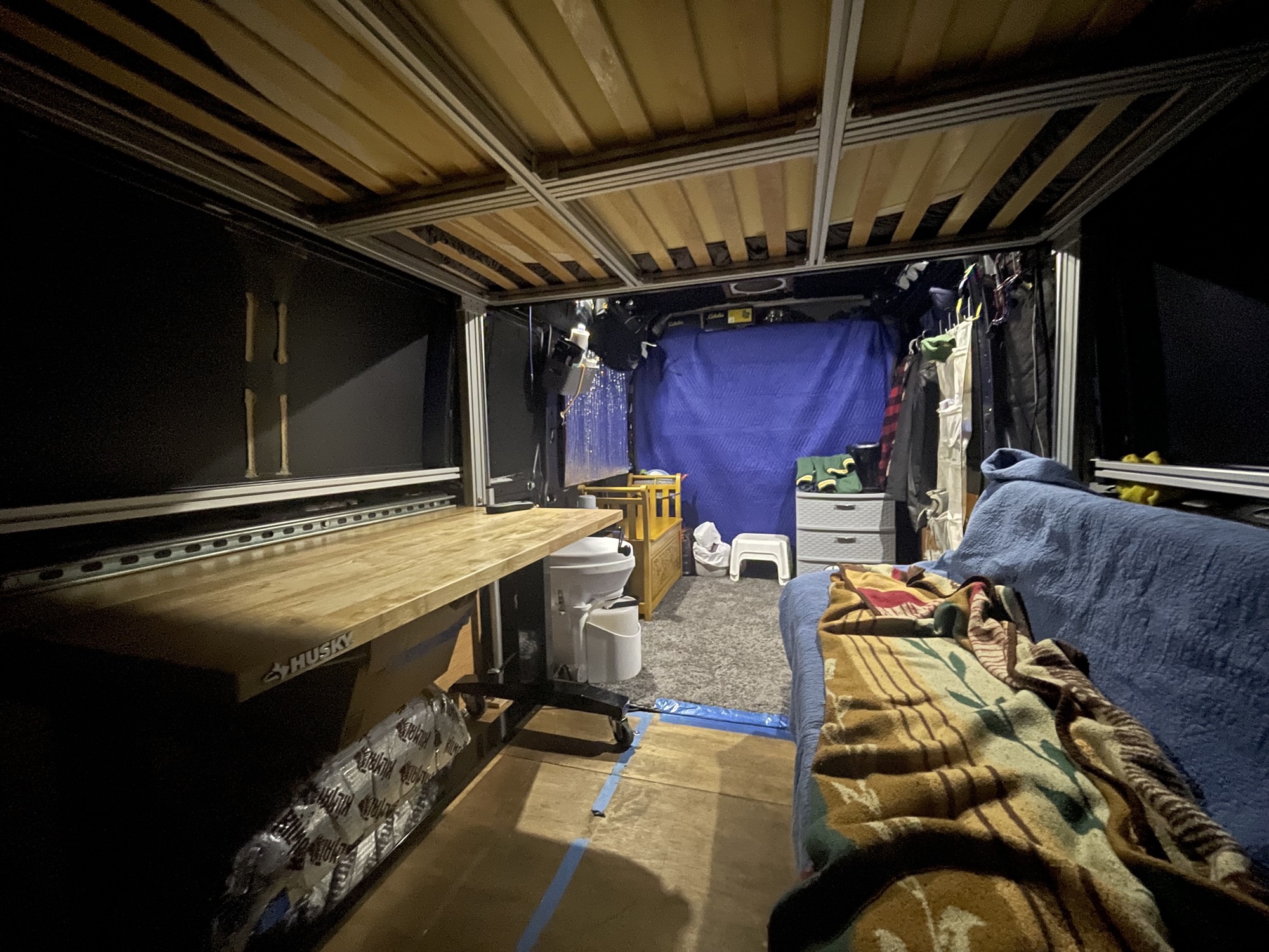 Inside the ProMaster at night: a plywood bed loft, a workbench, and a couch piled with blankets.
