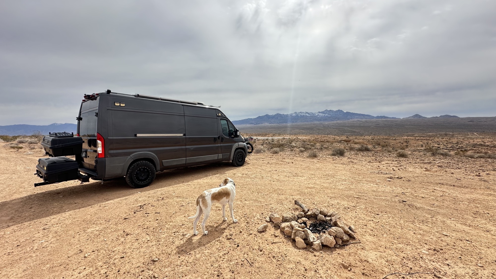Luca standing beside the ProMaster on open desert ground, with mountains on the horizon.